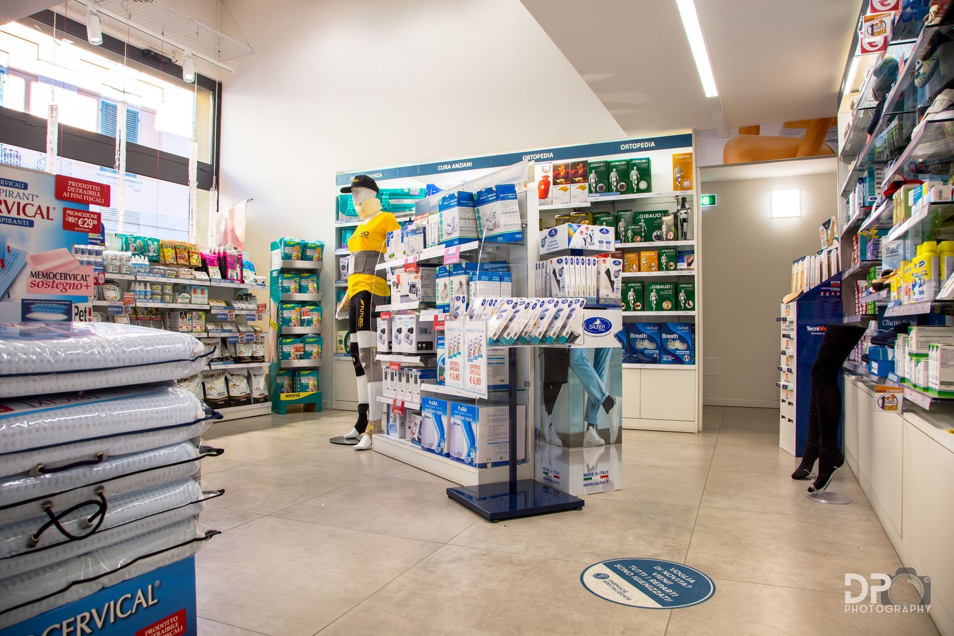 Interno di una farmacia ben illuminata, con corridoi dove sono esposti vari prodotti medici e sanitari sugli scaffali.