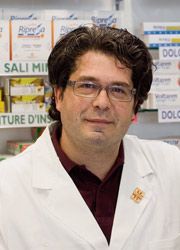 Un farmacista in camice bianco si trova in una farmacia, con scaffali pieni di scatole di medicinali sullo sfondo.