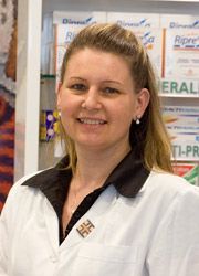 Una persona con indosso un camice bianco da laboratorio con colletto nero e un emblema, in piedi davanti agli scaffali dei prodotti farmaceutici.