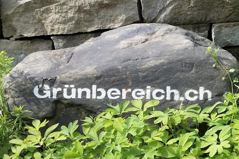 Der Text „Grünbereich.ch“ prangt auf einem dunklen Felsen inmitten grüner Pflanzen und einer Steinmauer.