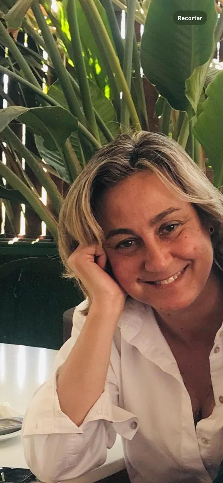 Mujer rubia sonríe, con la mano en la mejilla. Lleva una camisa blanca y está sentada al aire libre, cerca de plantas verdes.