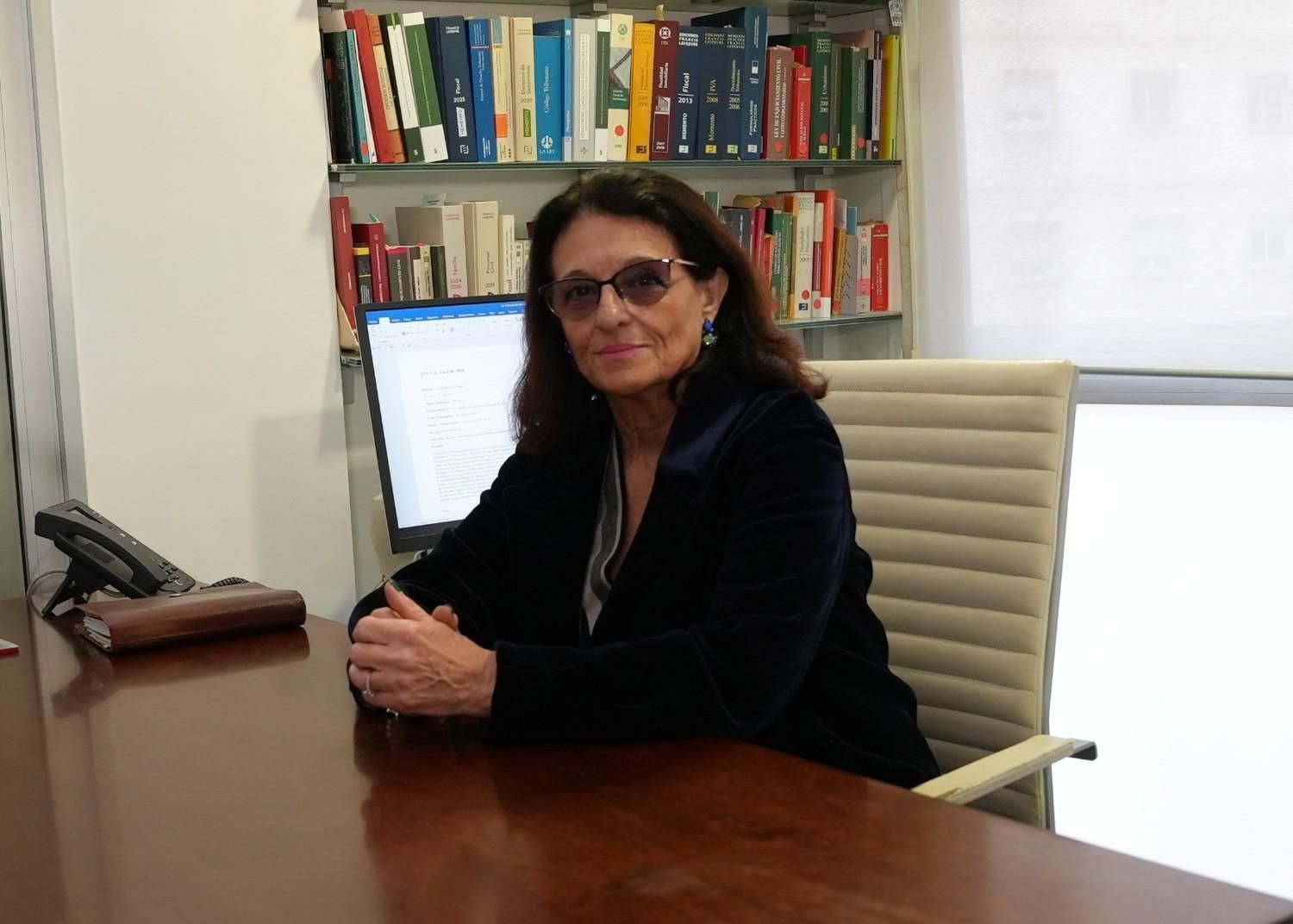 Mujer con gafas, sentada en un escritorio, con estanterías de libros al fondo.