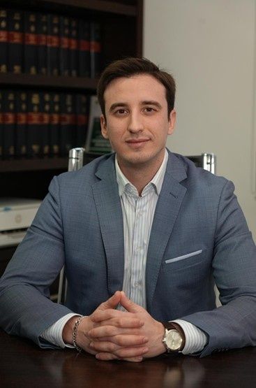 Hombre con una chaqueta azul sentado en un escritorio con las manos juntas, frente a una estantería.