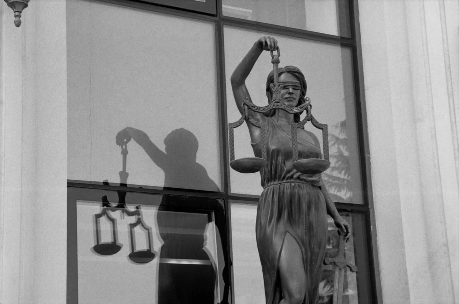 Estatua de la Dama de la Justicia sosteniendo una balanza, frente a una ventana, con sombra visible en el cristal.