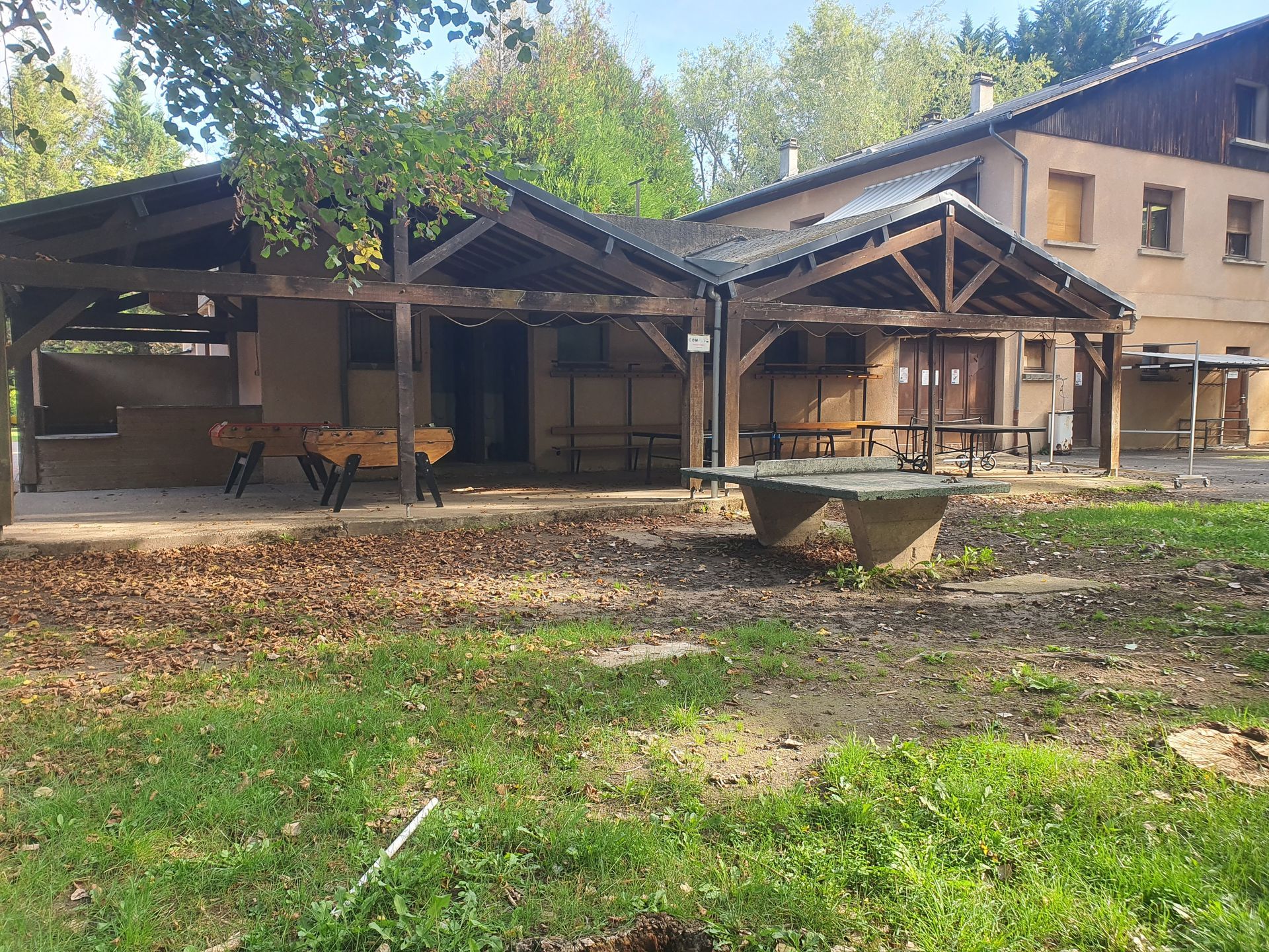 Chalet rustique avec une véranda ombragée, des poutres en bois et une jardinière en béton dans un jardin gazonné avec des arbres.