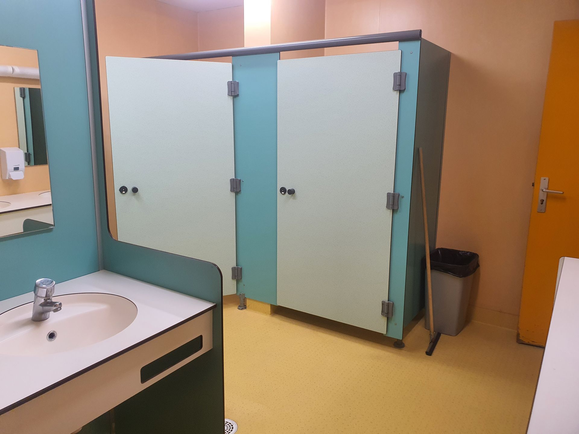 Toilettes avec lavabos, portes de cabine bleues, sol jaune et porte orange.
