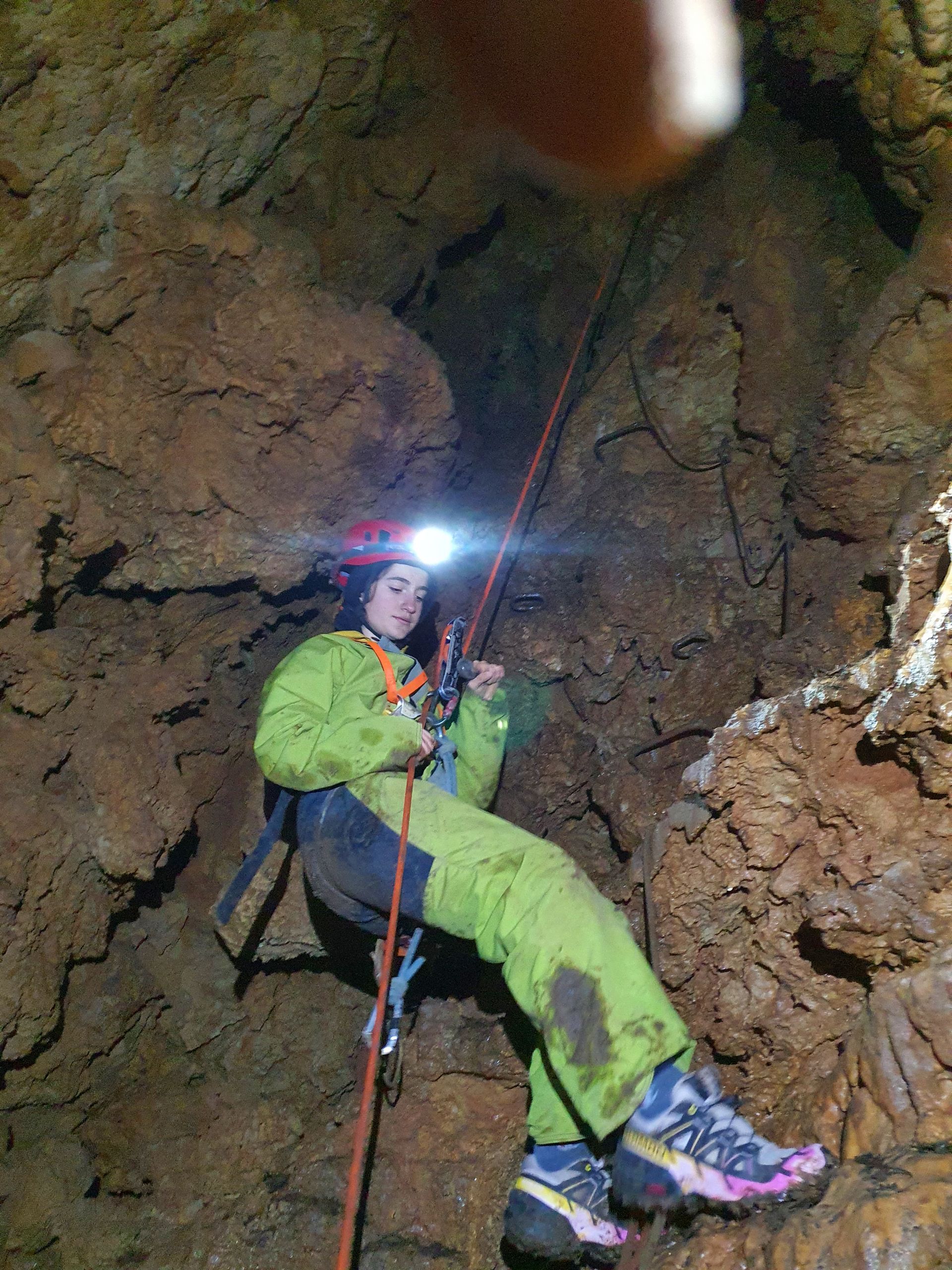 Un grimpeur en tenue verte descend en rappel à l'intérieur d'une grotte, sa lampe frontale éclairant la paroi rocheuse.