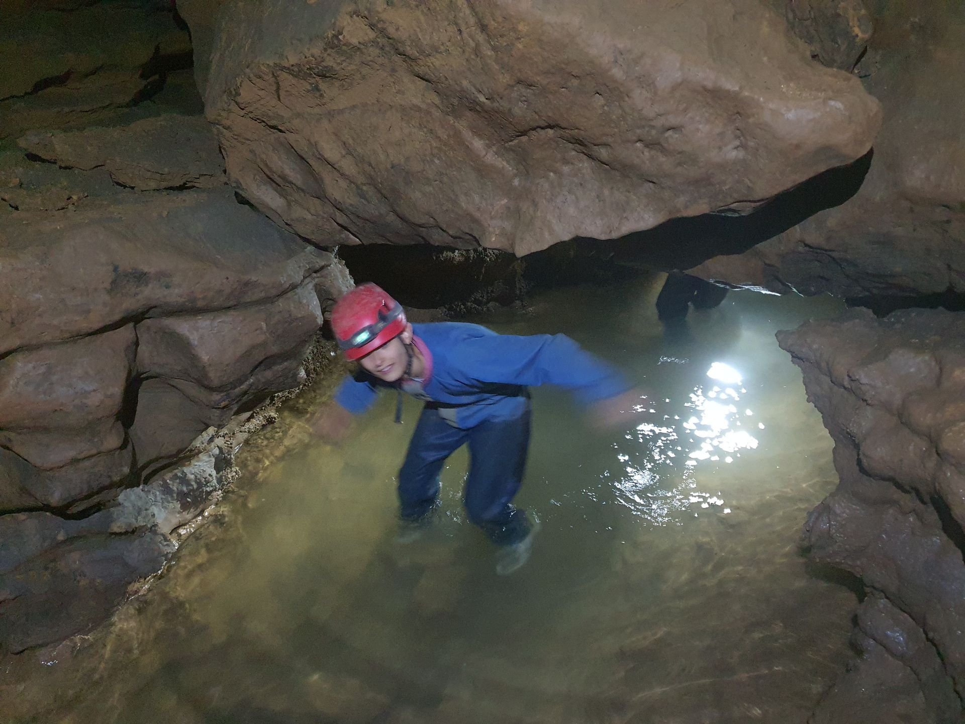 Un enfant portant un casque rouge se tient dans une mare peu profonde au pied d'imposantes formations rocheuses.