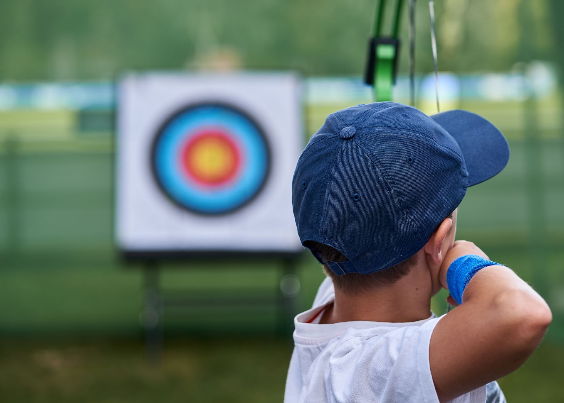 Enfant visant une cible de tir à l'arc en extérieur, vu de dos, portant une casquette et un t-shirt bleus.