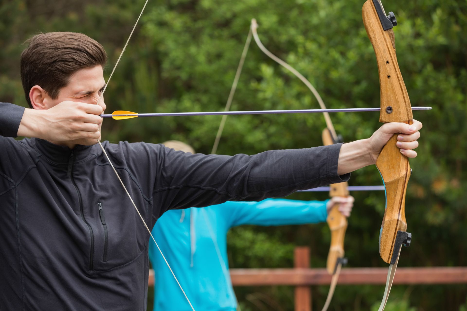 Un homme bande un arc en bois à l'extérieur, visant une flèche avec une expression concentrée.