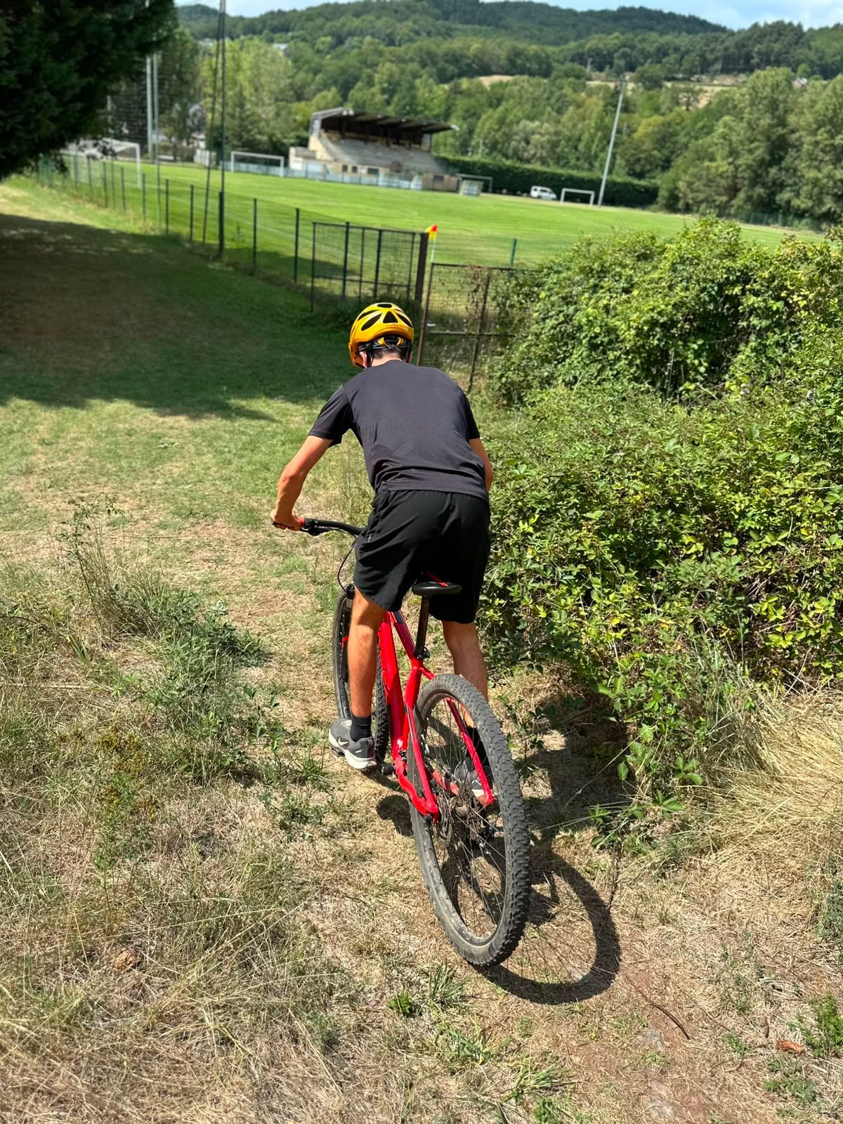 Cycliste en maillot noir et casque jaune, à vélo de montagne rouge, sur un chemin herbeux longeant une clôture et des champs.