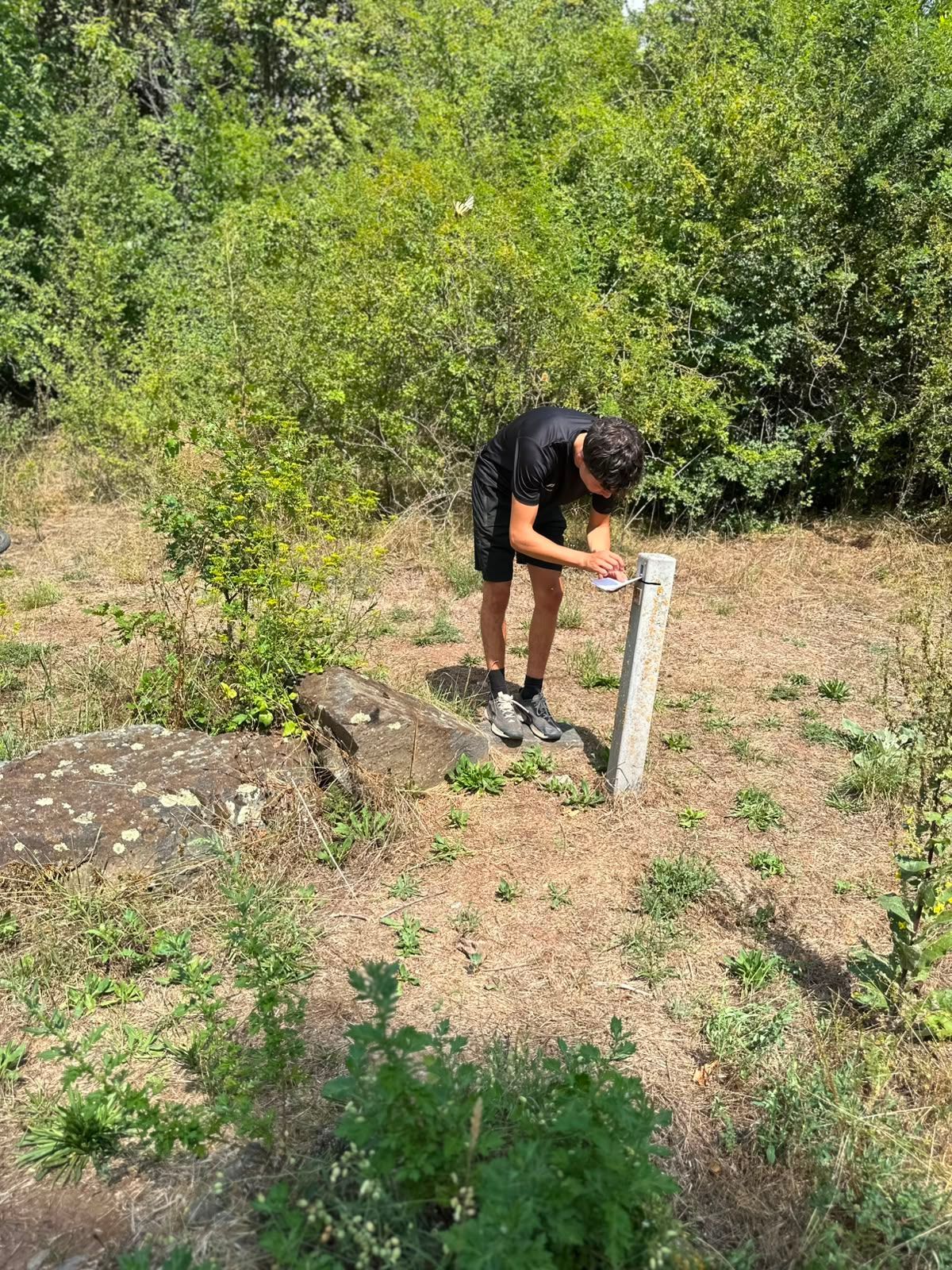 Une personne penchée sur un poteau blanc dans un champ herbeux bordé d'arbustes.