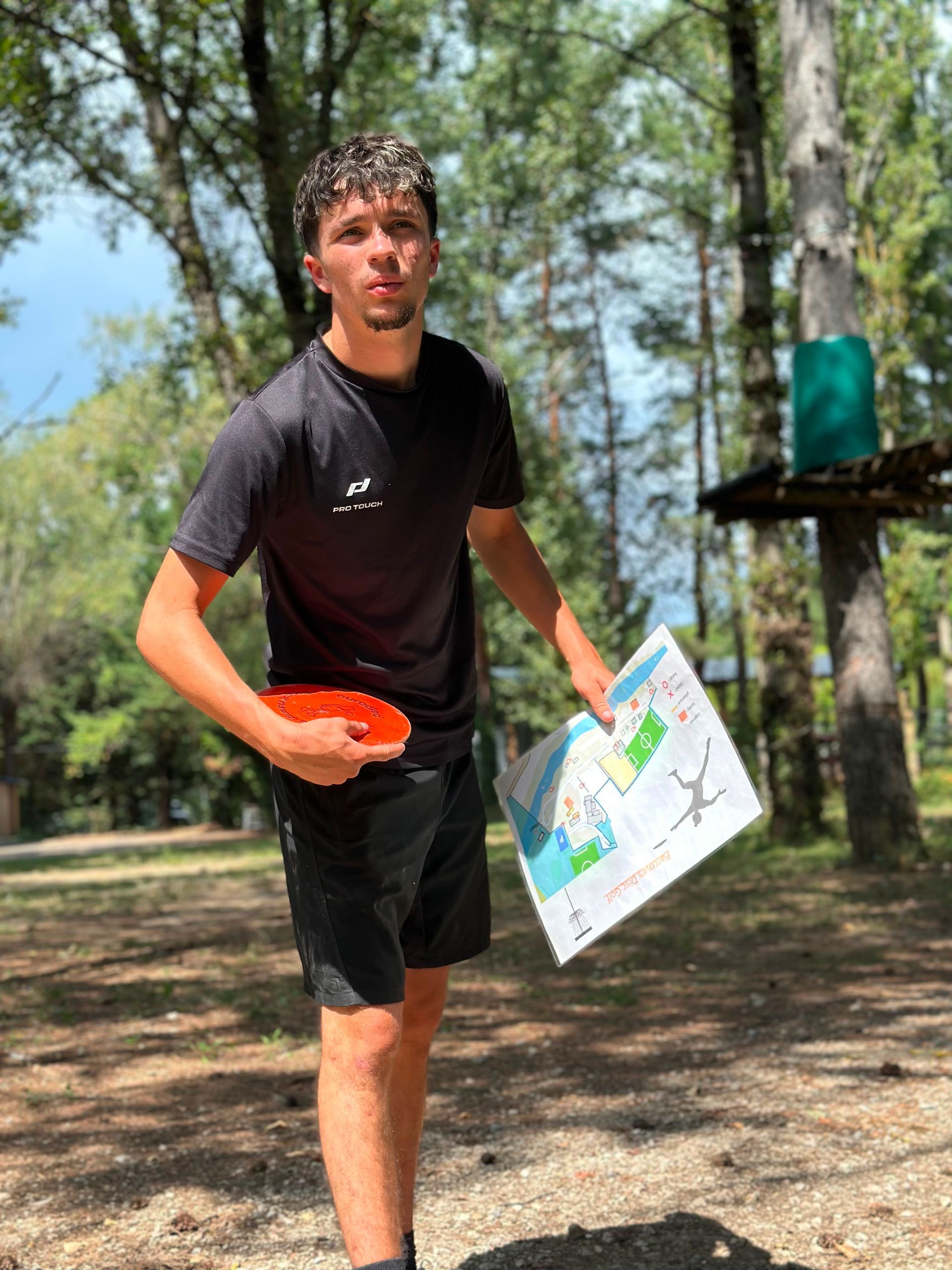 Un homme tenant une carte et un frisbee orange se promène sur un sentier boisé, en plein soleil.