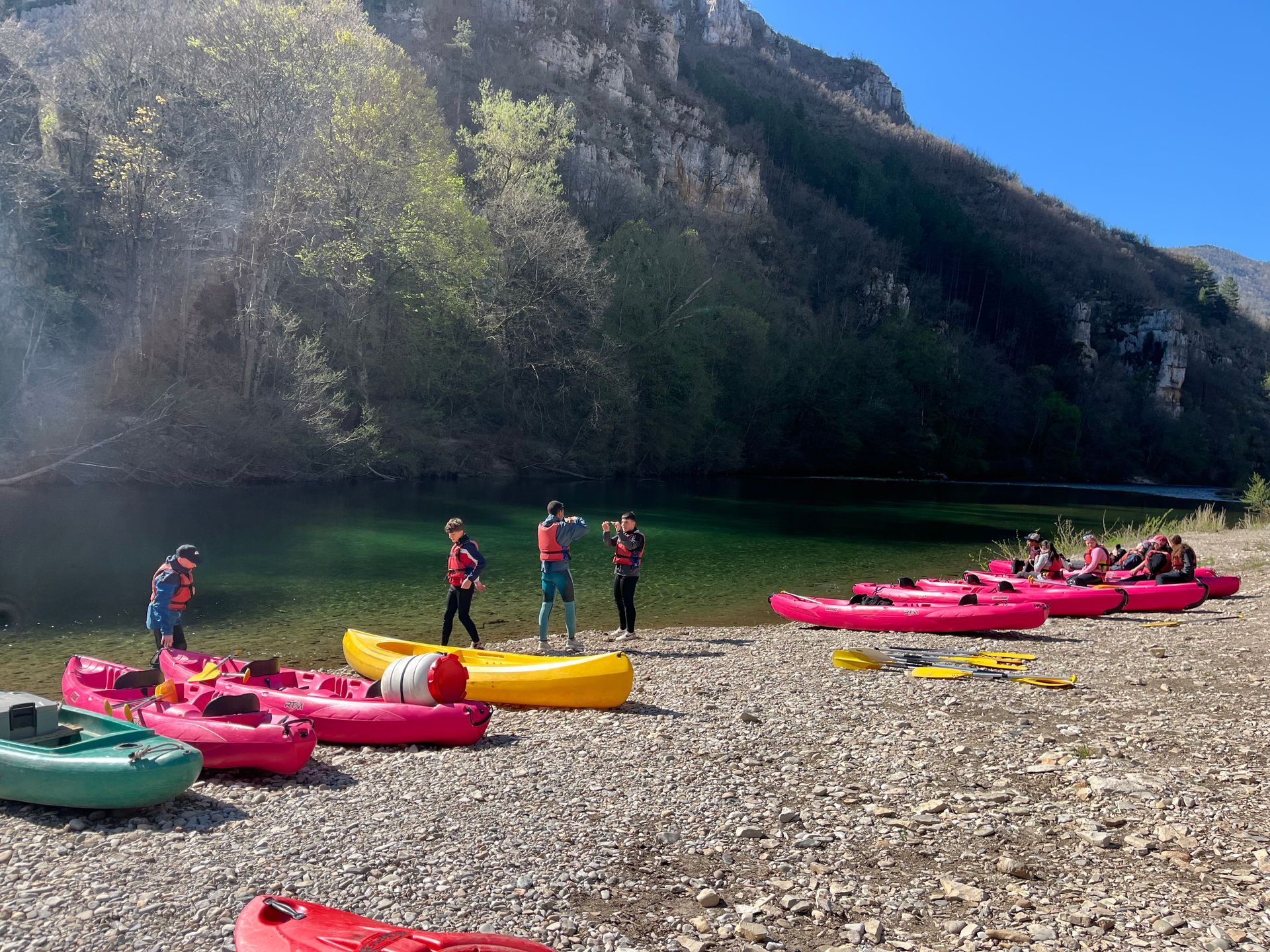 Des personnes en gilets de sauvetage rouges préparent des radeaux colorés sur une rive rocheuse, au bord d'une rivière calme, avec les montagnes pour toile de fond.