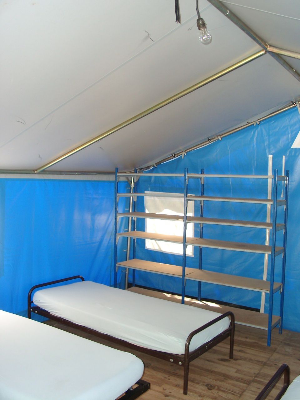 Deux lits de camp simples à l'intérieur d'une tente bleue ou d'un abri temporaire avec des étagères métalliques sur la droite.