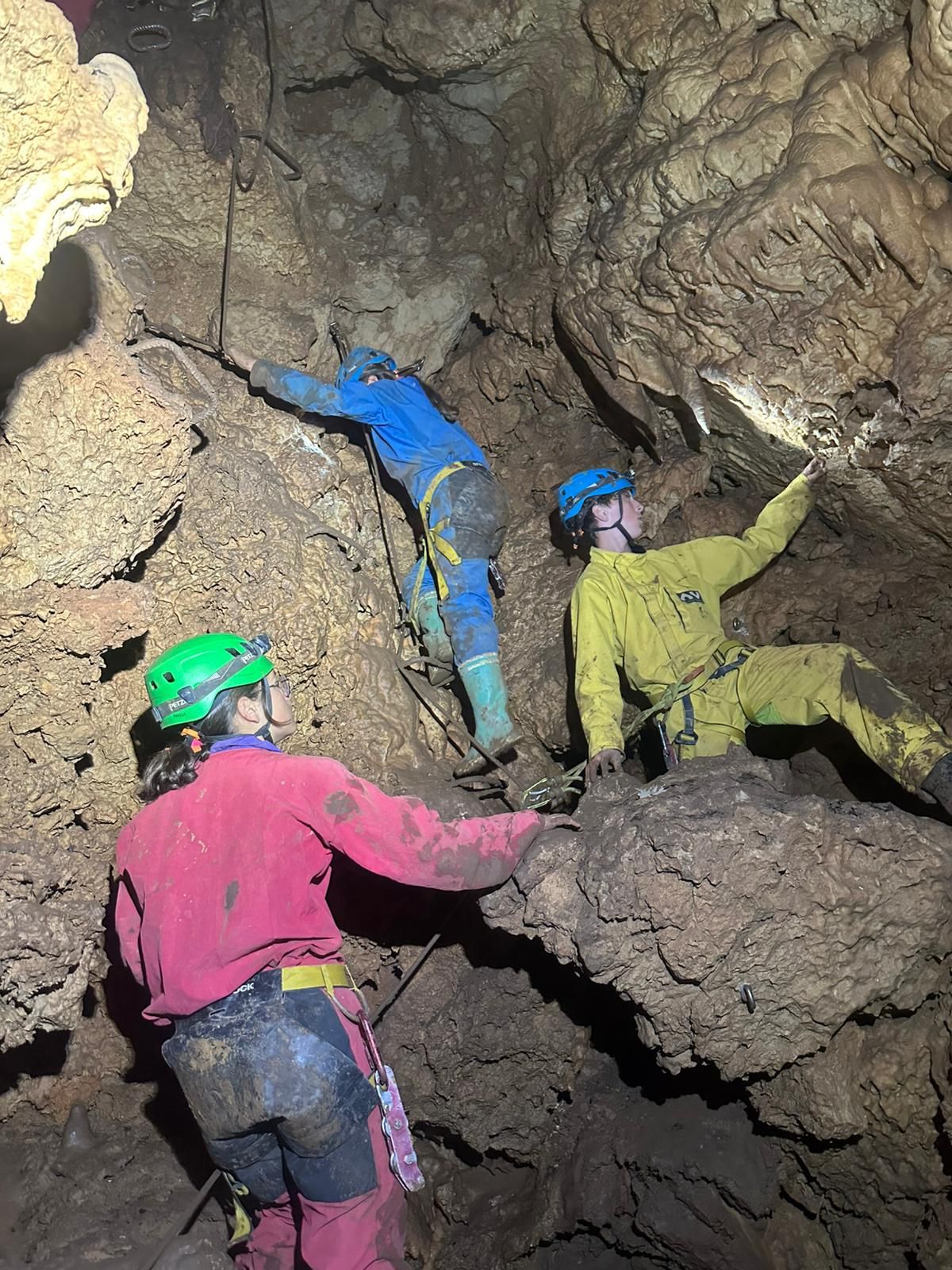 Trois spéléologues casqués escaladent un étroit passage rocheux dans une grotte.