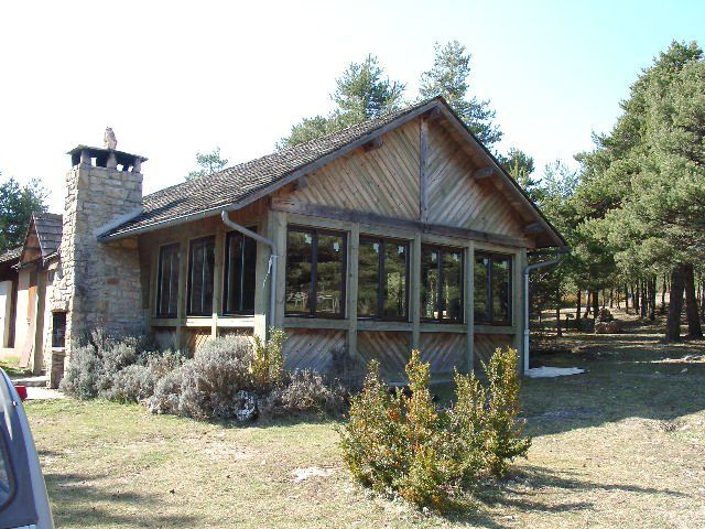 Maison de style chalet avec cheminée en pierre et grandes fenêtres dans une cour boisée et herbeuse