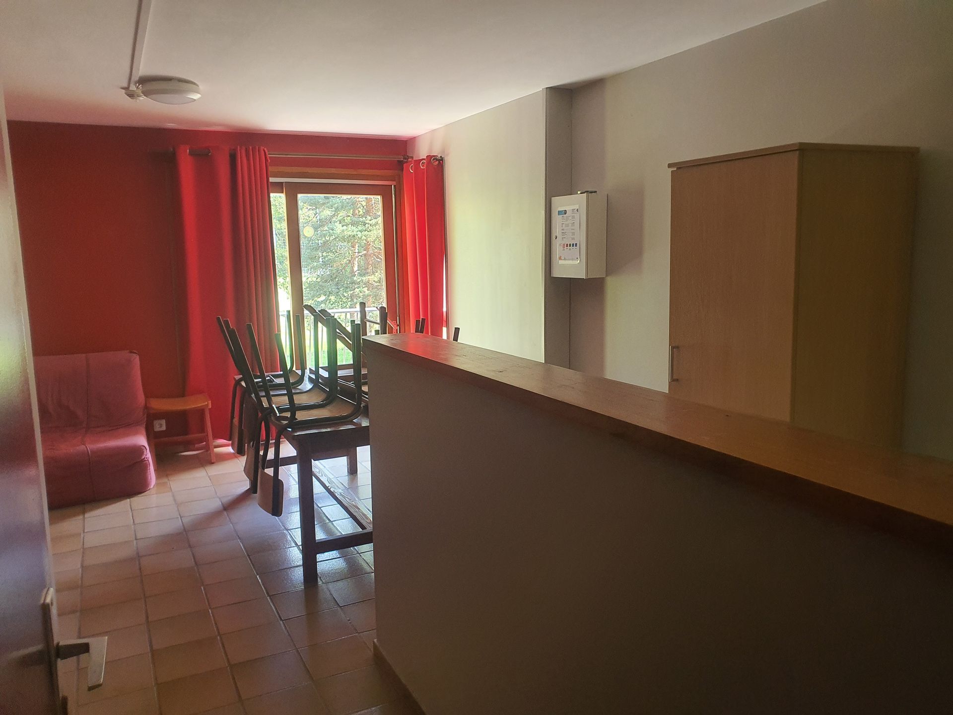 Petit salon avec rideaux rouges, sol carrelé, chaises et une table près d'une fenêtre.