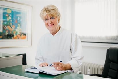 Eine Frau im weißen Kittel sitzt mit einem Buch an einem Schreibtisch.