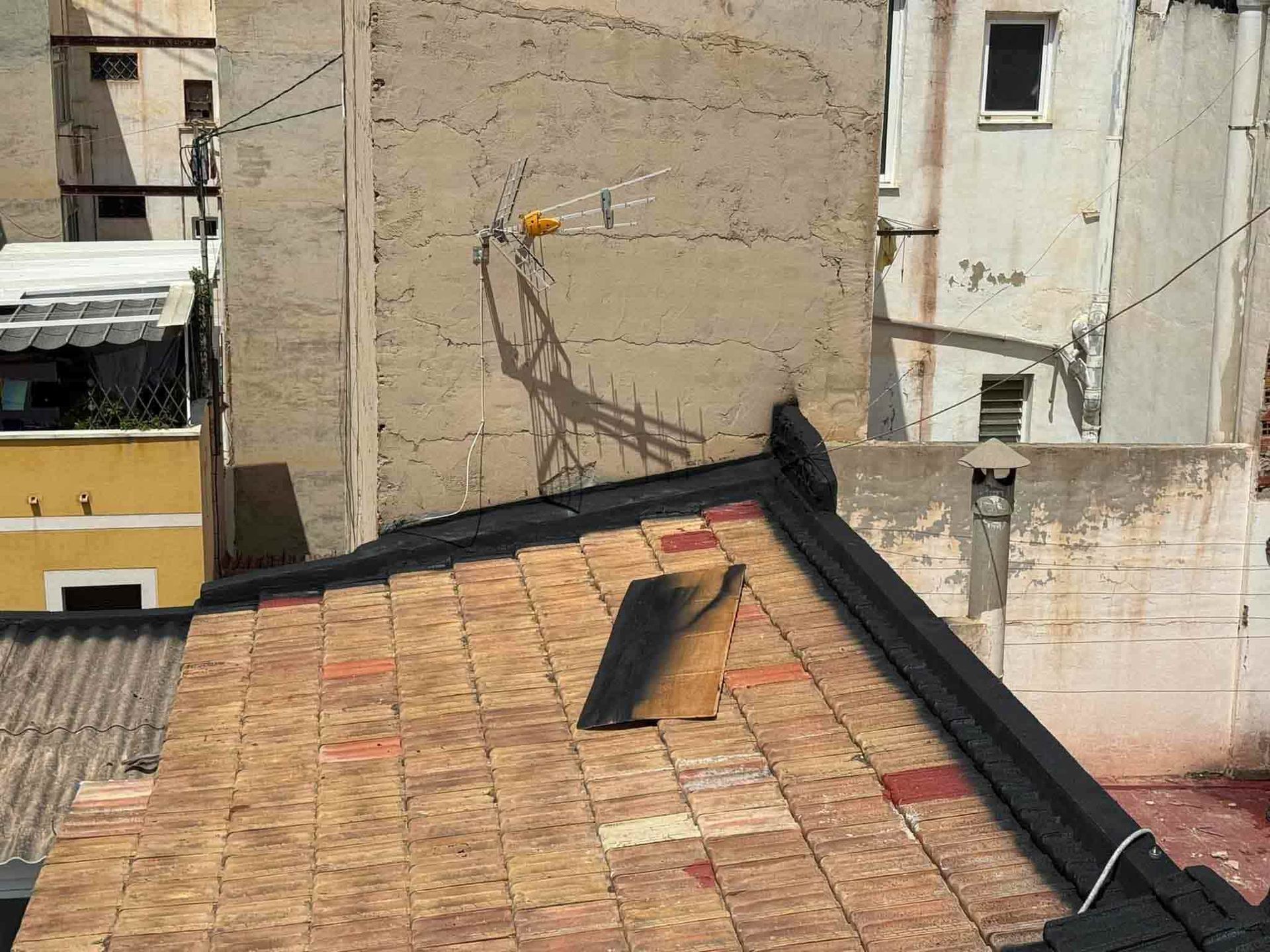 Vista de la azotea con tejas de ladrillo, sombra de antena en una pared de hormigón y otros edificios al fondo.