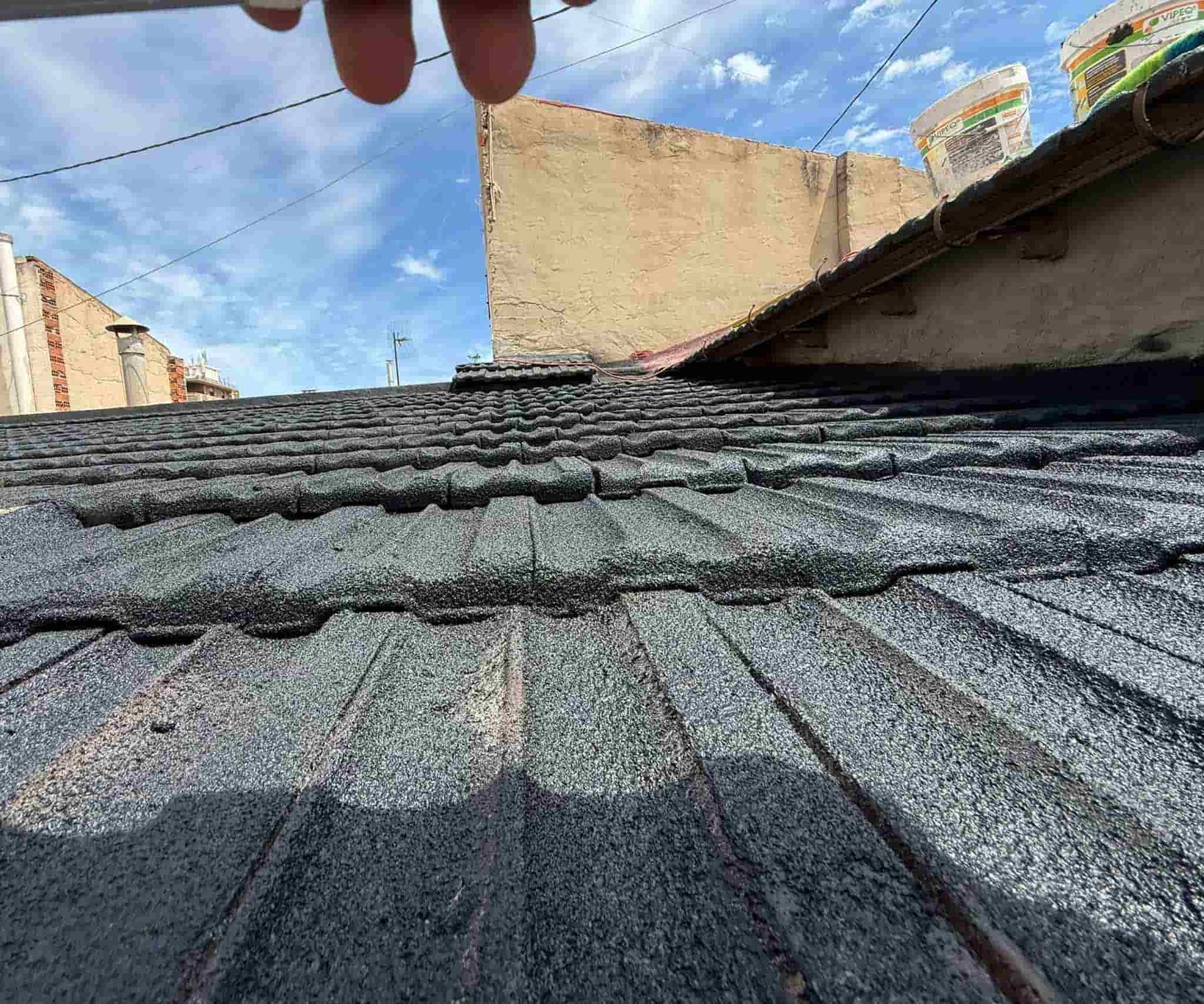 Primer plano de un techo negro texturizado con una mano encima contra un cielo azul nublado, edificios en el fondo.