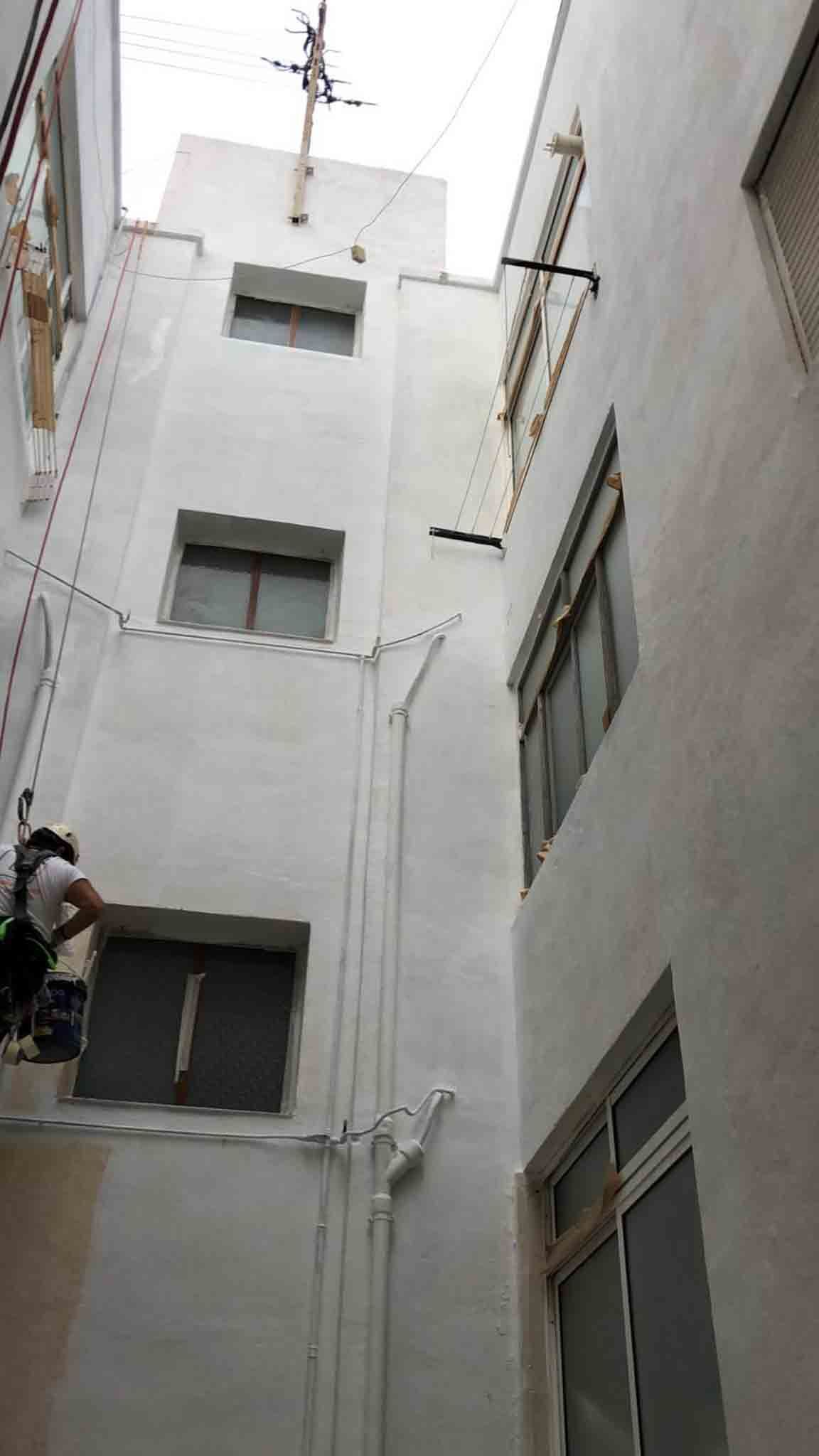 Callejón estrecho entre edificios blancos; trabajador pintando una ventana.