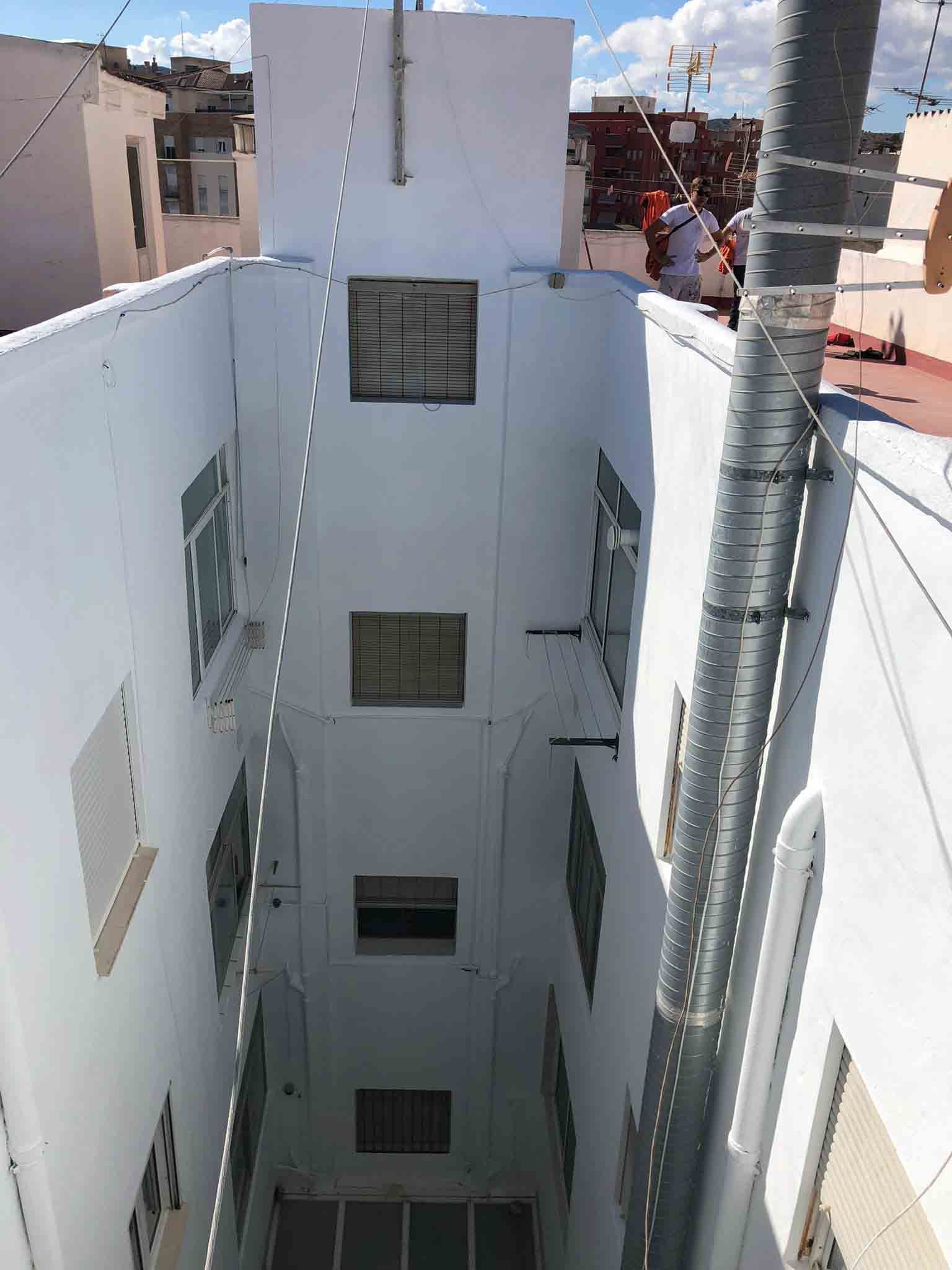 Hueco estrecho de paredes blancas con múltiples ventanas. Tubería metálica y tejado a la derecha.