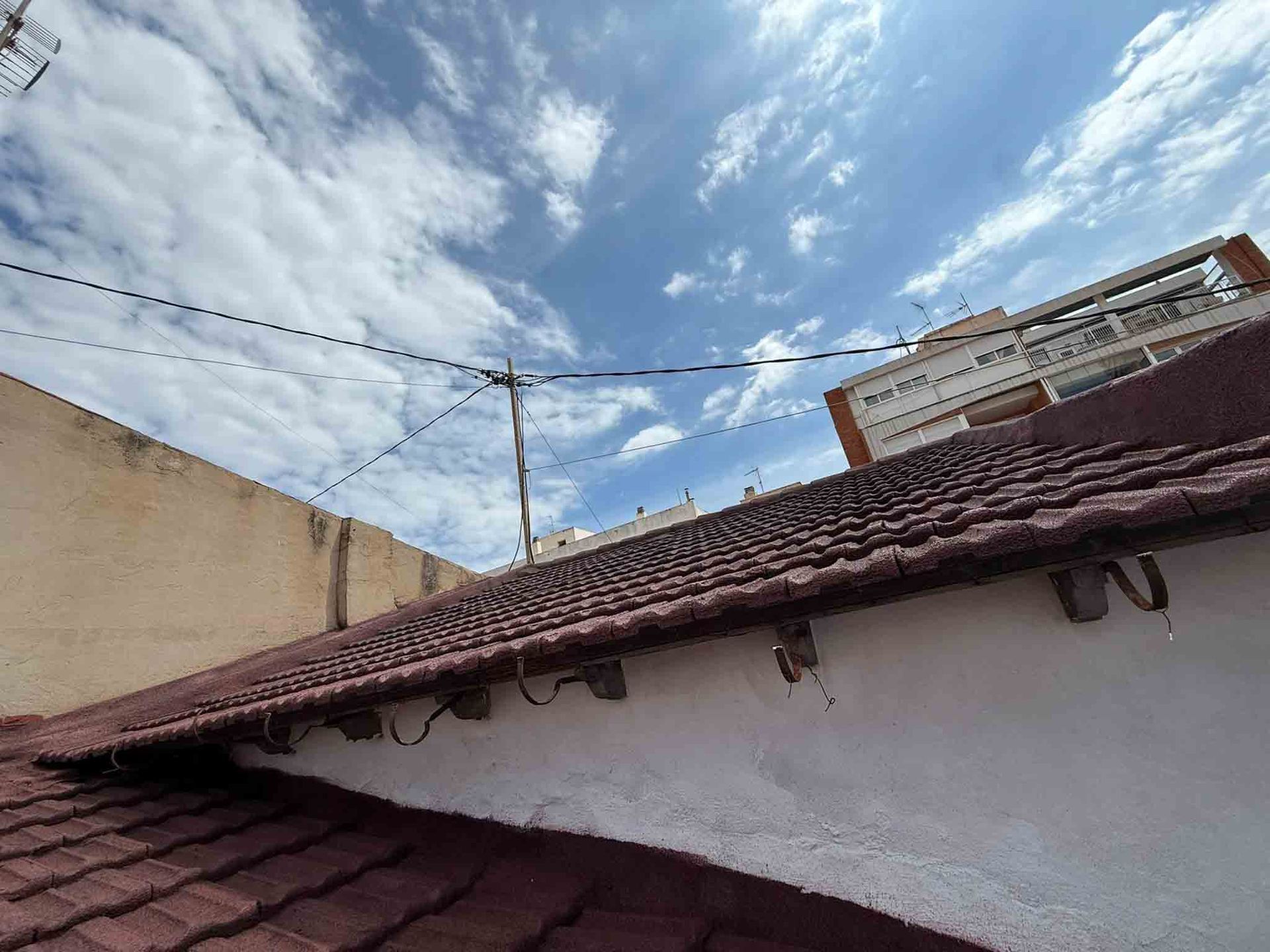 Azotea con tejas rojas contra un cielo azul brillante con nubes dispersas; se ven líneas eléctricas y un edificio.