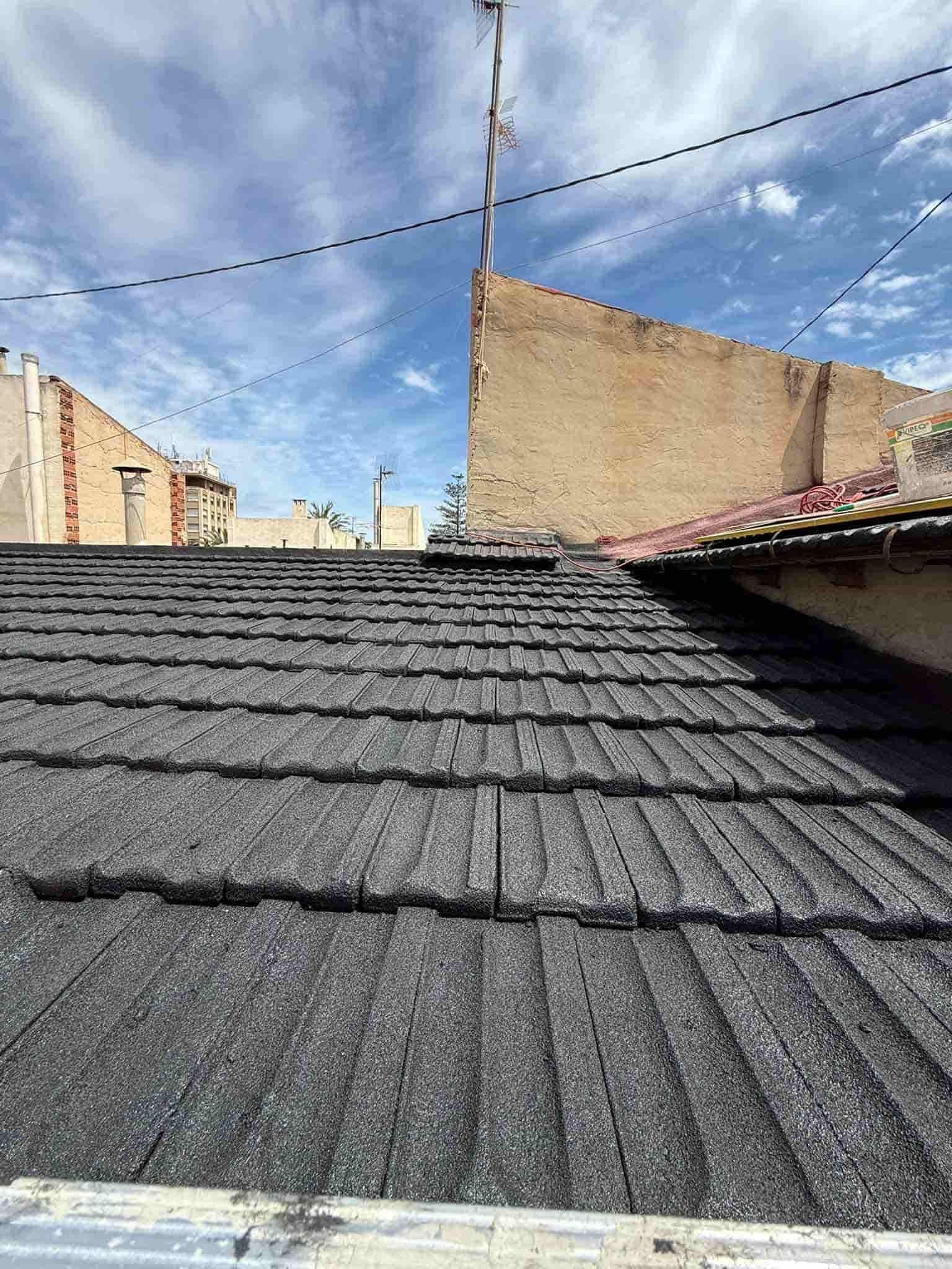 Tejas de asfalto negro sobre un tejado, con una pared de ladrillos al fondo bajo un cielo azul.