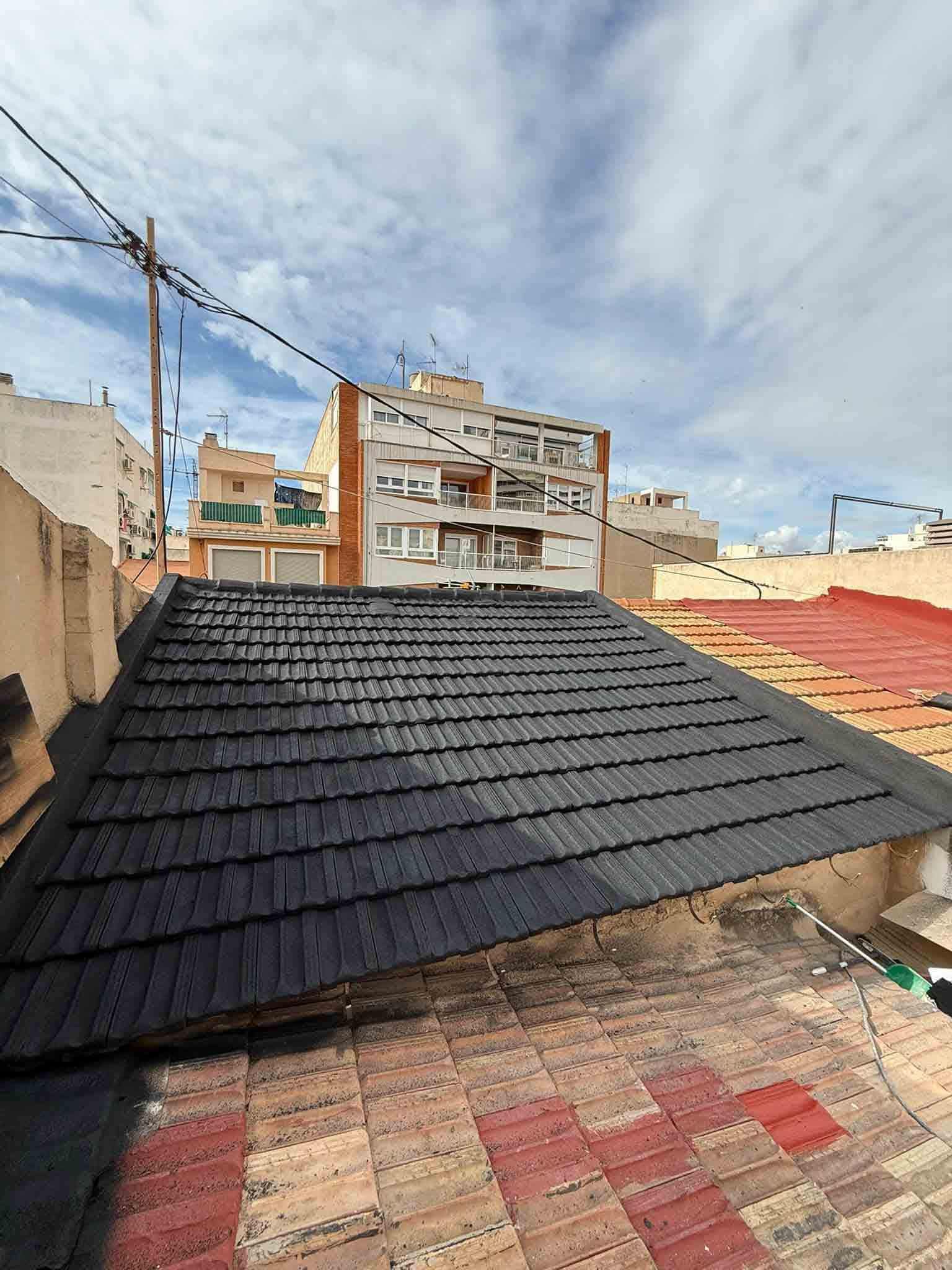 Vista de un tejado con tejas negras. Al fondo, edificios y un cielo nublado.