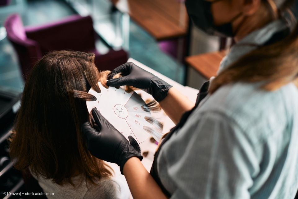 Eine Person mit schwarzen Handschuhen hält eine Haarprobe an eine Farbkarte. Eine Person mit Maske schaut zu.