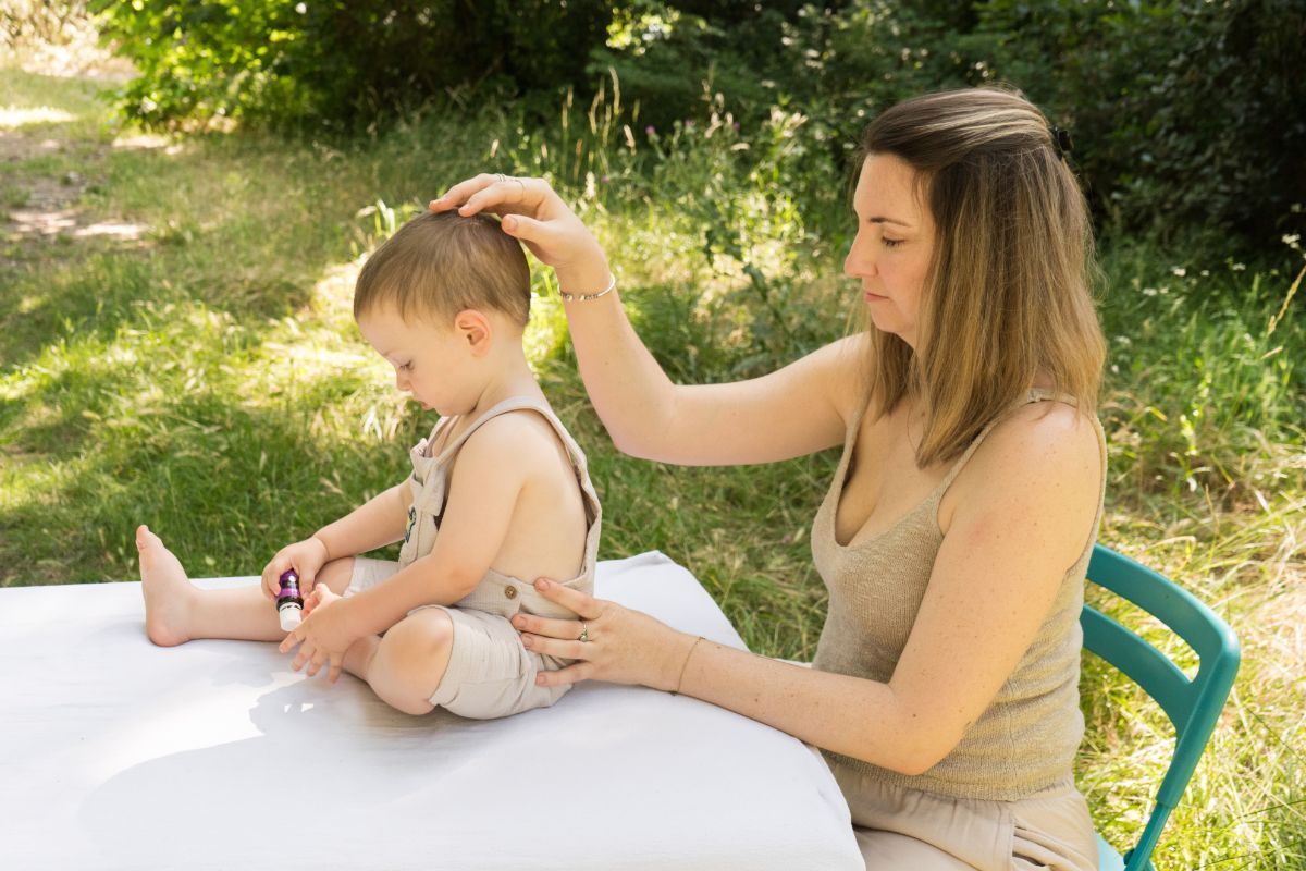 Una mujer cepilla el cabello de su hijo pequeño al aire libre, sentada a una mesa blanca en un jardín soleado.