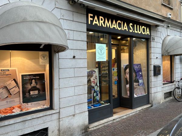Farmacista in camice bianco sorride, braccia incrociate, in una farmacia con scaffali pieni di medicinali.