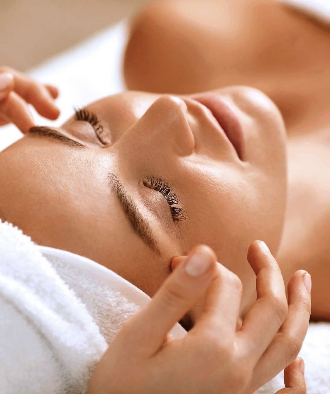 Una persona, con los ojos cerrados, recibe un suave masaje facial mientras está tumbada con una toalla blanca