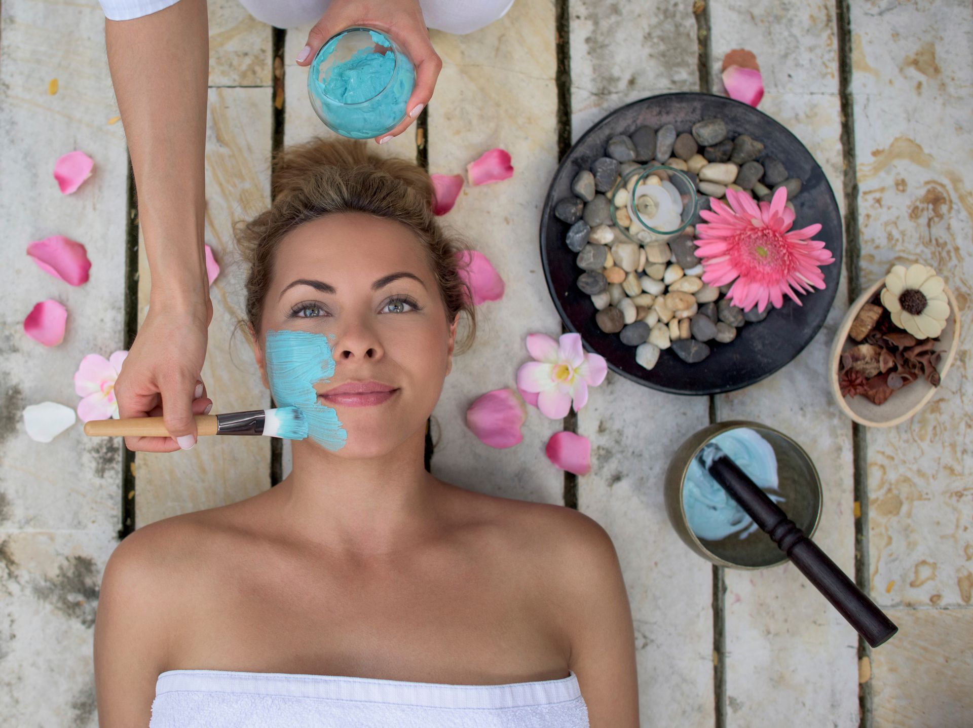 Una persona recibe la aplicación de una mascarilla facial azul en un spa, rodeada de pétalos de flores
