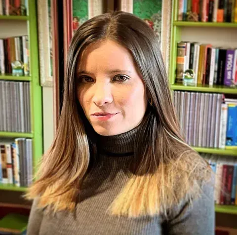 Mujer con cabello largo castaño, vistiendo un suéter de cuello alto gris, estanterías en el fondo.