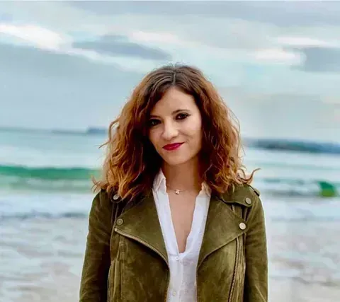 Mujer con chaqueta verde oliva y camisa blanca, sonriendo, de pie en una playa con cielo nublado.