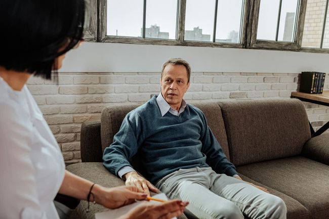 Mujer rubia con blazer blanco, mirando hacia abajo. Interior con mesa.