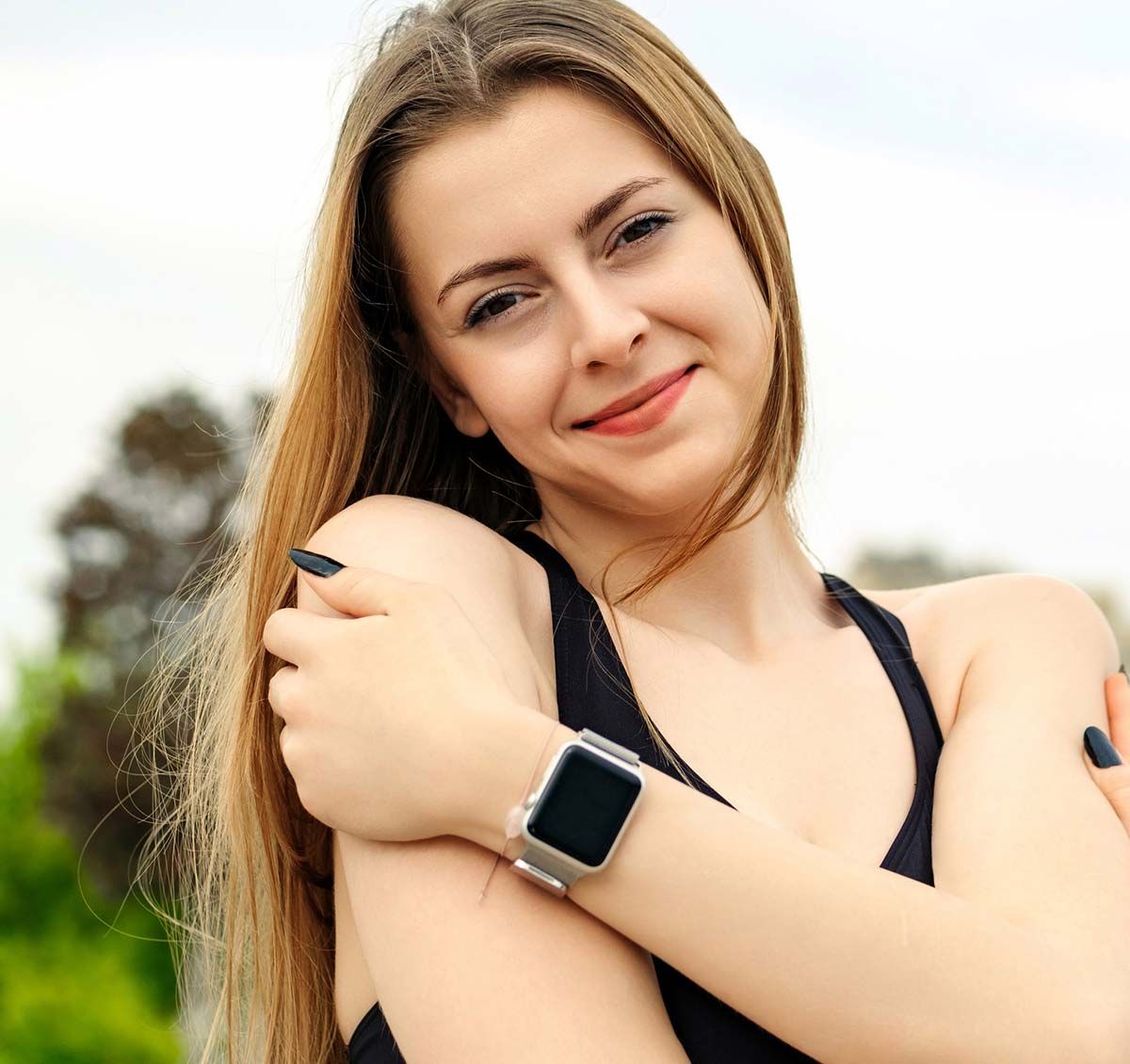 Mujer con cabello rubio largo, vestida con una blusa negra y un reloj inteligente, sonríe mientras se abraza a sí misma al aire libre.