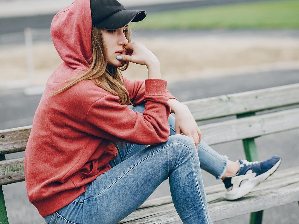 Mujer con sudadera roja con capucha, vaqueros y gorra negra, sentada en un banco de madera, con aspecto pensativo.