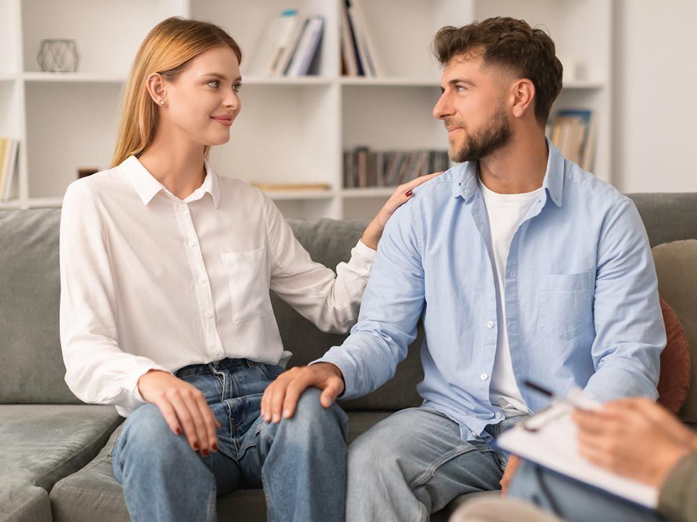 Pareja sentada en un sofá, la mano de la mujer sobre el hombro del hombre. Se miran el uno al otro; en primer plano, la terapeuta con su portapapeles.