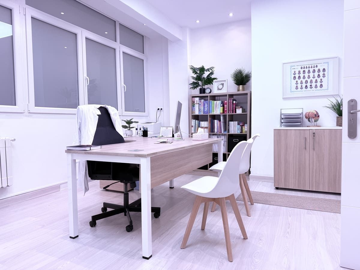 Oficina luminosa con escritorio, sillas, estantería y armario. Ventanas al fondo.