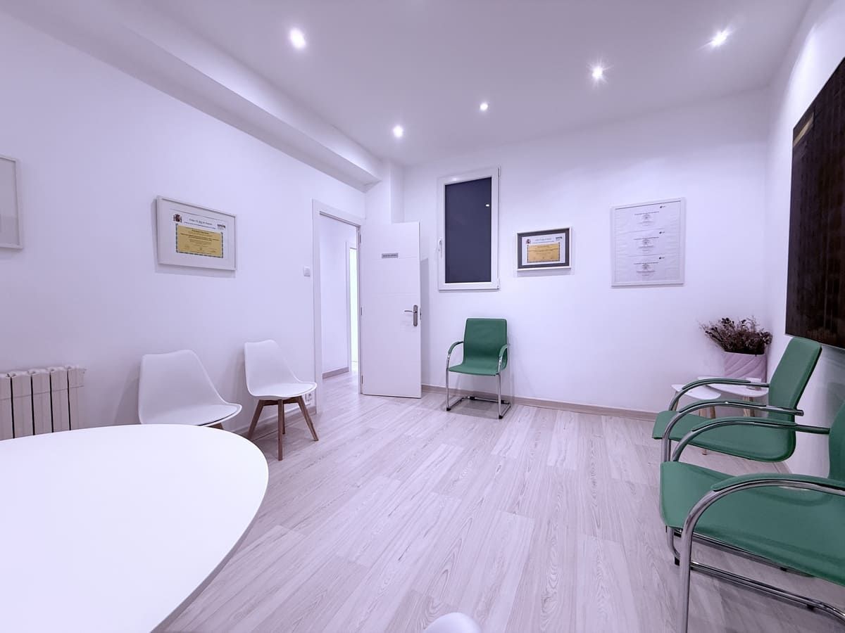 Sala de espera con paredes blancas, sillas y una mesa redonda. Sillas verdes y blancas. Puerta abierta.