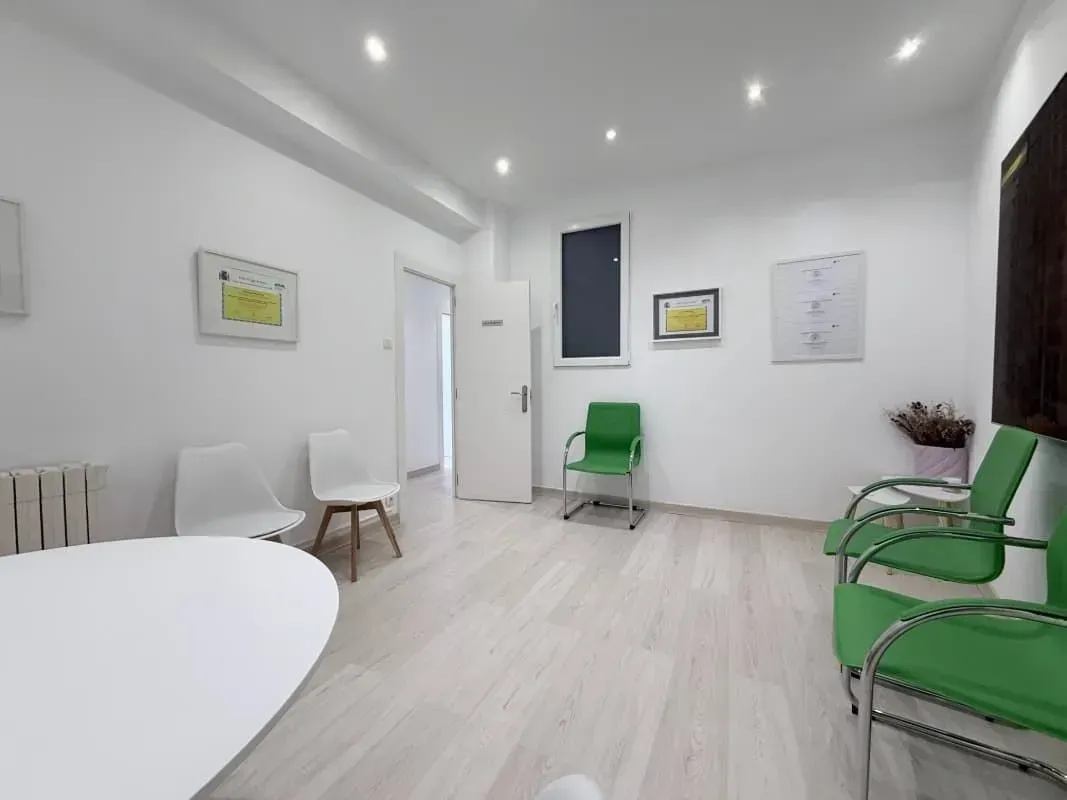 Sala de espera luminosa con paredes blancas, suelo de madera clara, sillas blancas y sillones verdes junto a obras enmarcadas.