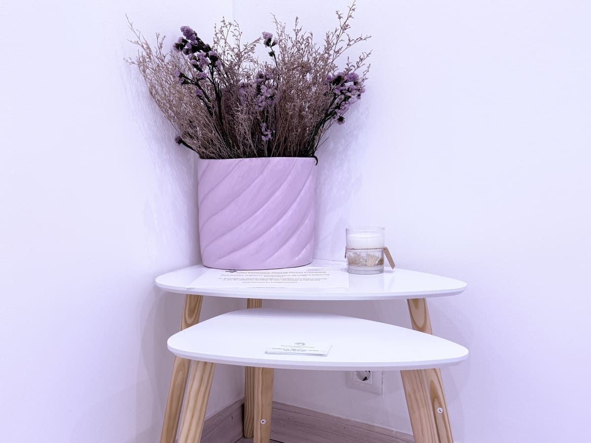 Mesitas auxiliares blancas apilables en la esquina; jarrón morado con flores secas; vela.