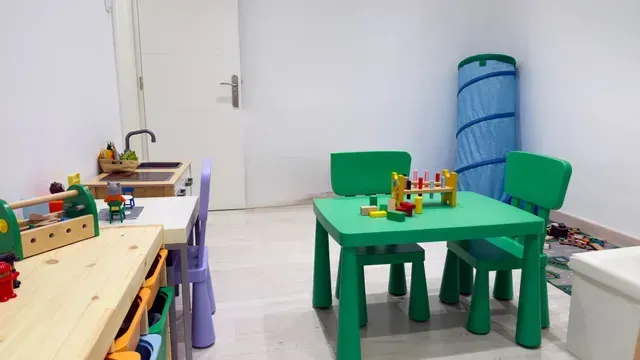 Sala de juegos luminosa con mesa y sillas de juguete verdes, cocinita de juguete de madera y juguetes coloridos esparcidos por todas partes.
