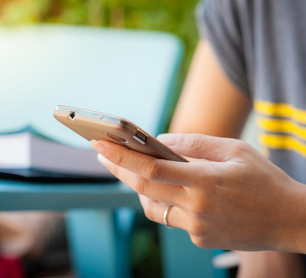 Persona sosteniendo un teléfono inteligente dorado, al aire libre, con un libro y una silla color verde azulado al fondo.