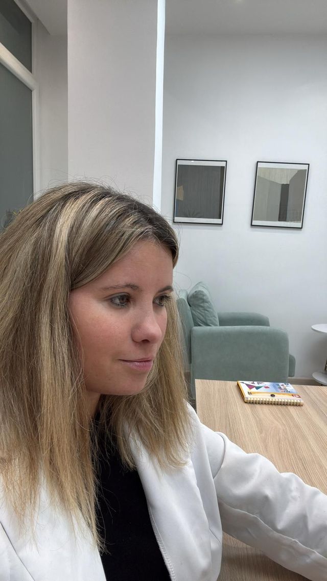 Mujer rubia con blazer blanco, mirando hacia abajo. Interior con mesa.