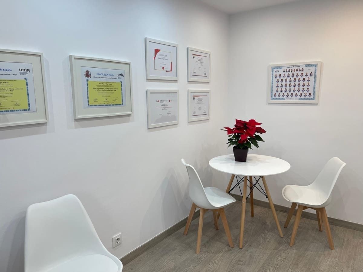 Zona de espera con sillas y mesa blancas, certificados enmarcados en la pared, planta en maceta.