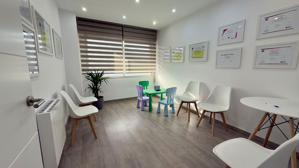Una luminosa sala de espera con varias sillas blancas, una pequeña mesa colorida para niños y documentos enmarcados en las paredes.